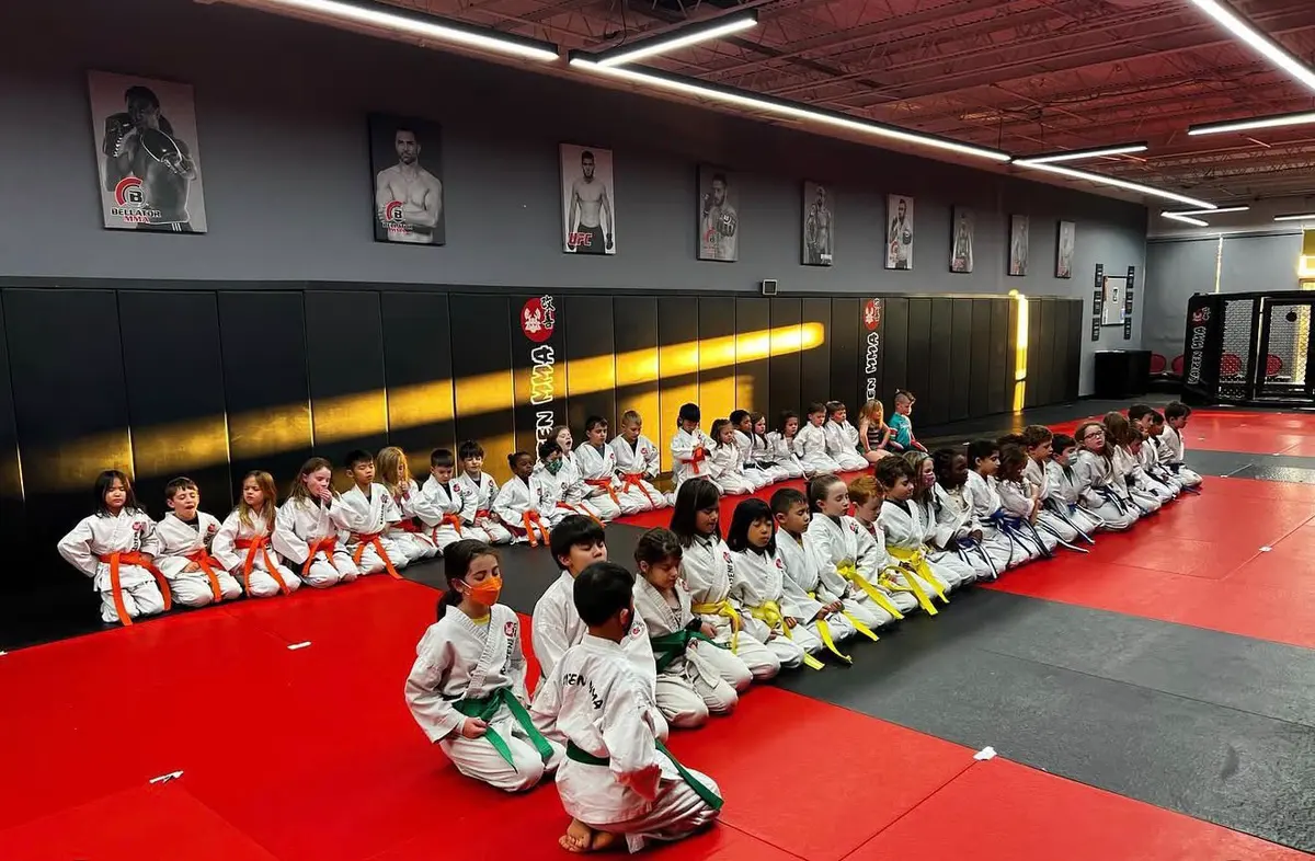 Children in martial arts uniforms sitting along the edge of a training mat